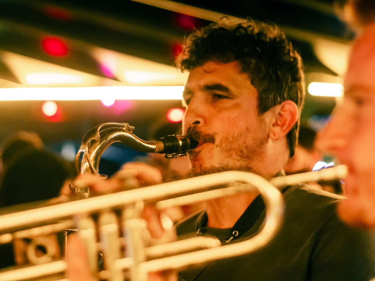 Saxofonista sopra o sax em close, com o metal dourado de um trompete em primeiro plano