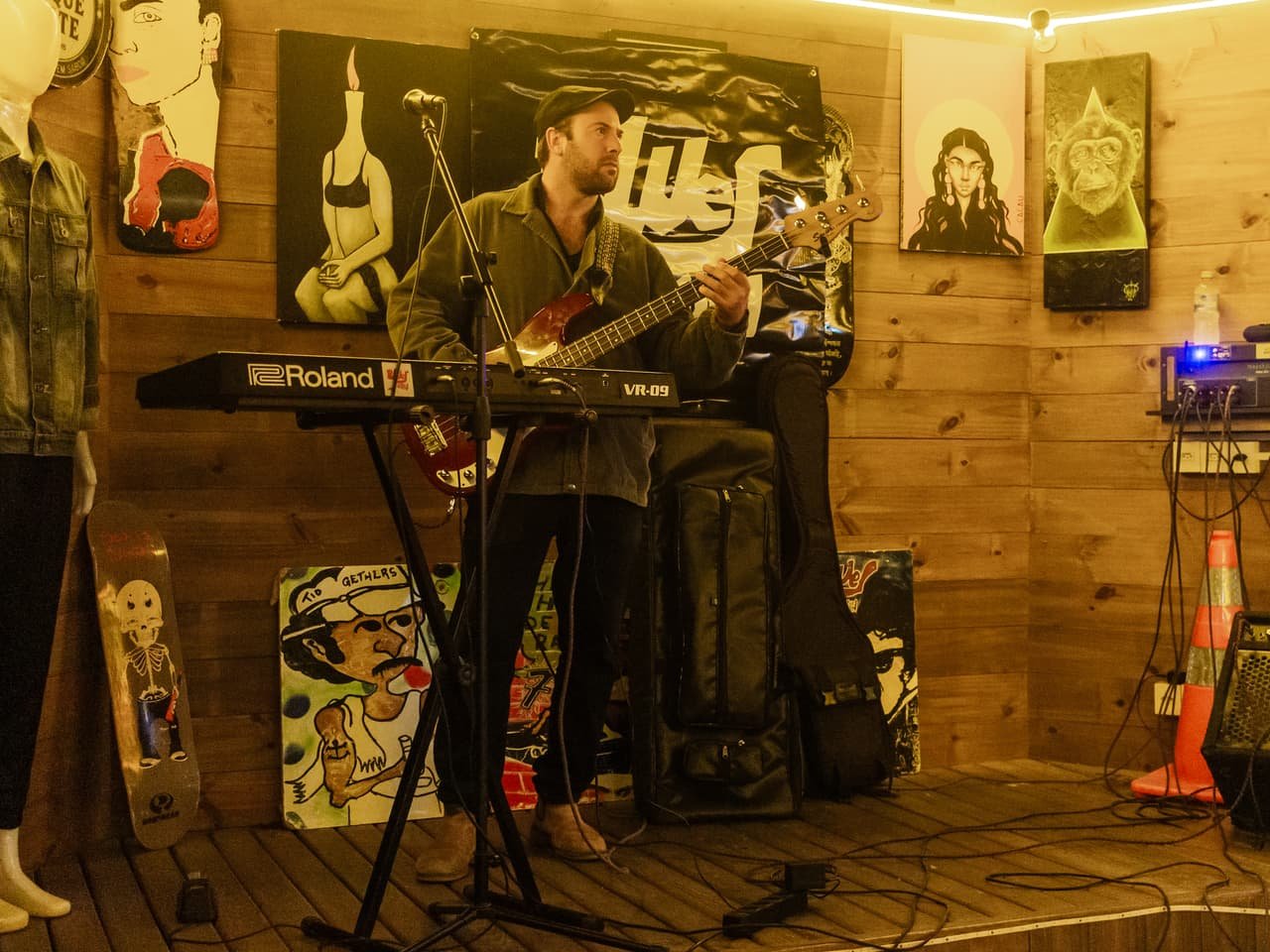 Baixista canta atrás de um teclado Roland num canto de palco de madeira decorado com quadros e skates
