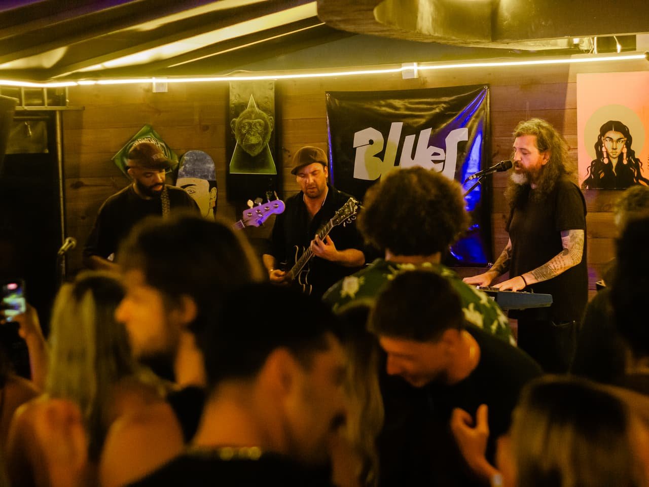 Banda toca sob luz quente diante do banner Blues Session enquanto o público lotado ocupa a frente do palco