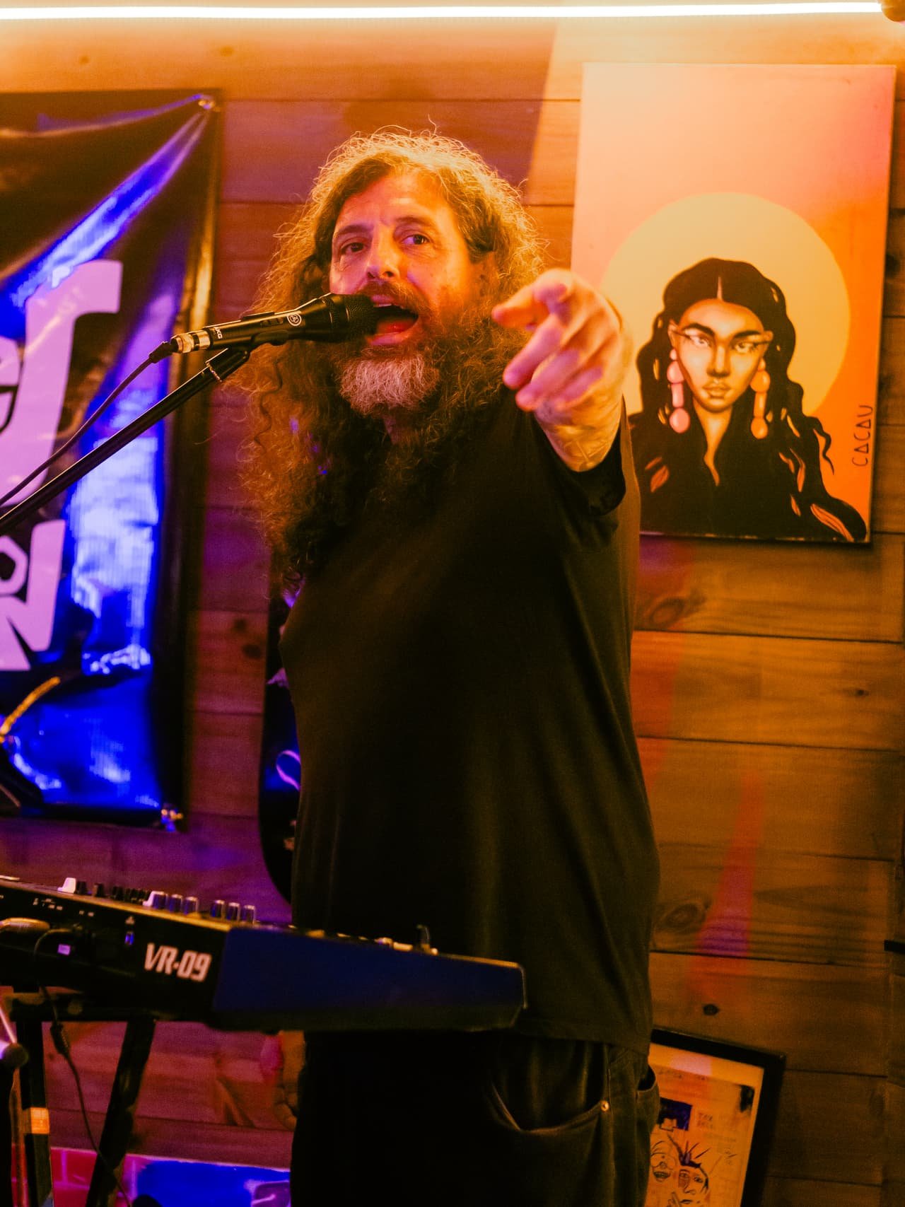 Cantor de barba e cabelos longos canta ao microfone apontando para o público, ao lado de um teclado