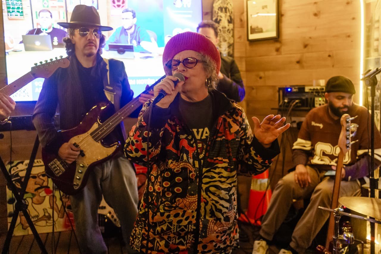 Cantora de chapéu vermelho e roupa estampada canta acompanhada por baixista de chapéu e baterista