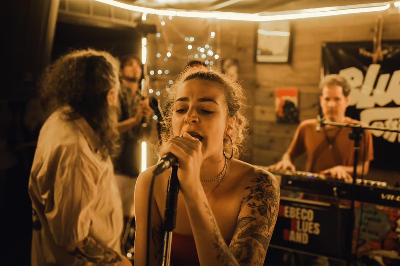 Cantora de cabelos cacheados canta de olhos fechados ao microfone enquanto o tecladista toca atrás dela