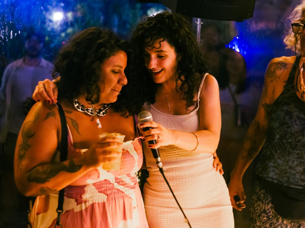 Duas mulheres abraçadas dividem o microfone durante a jam, sob luz azulada