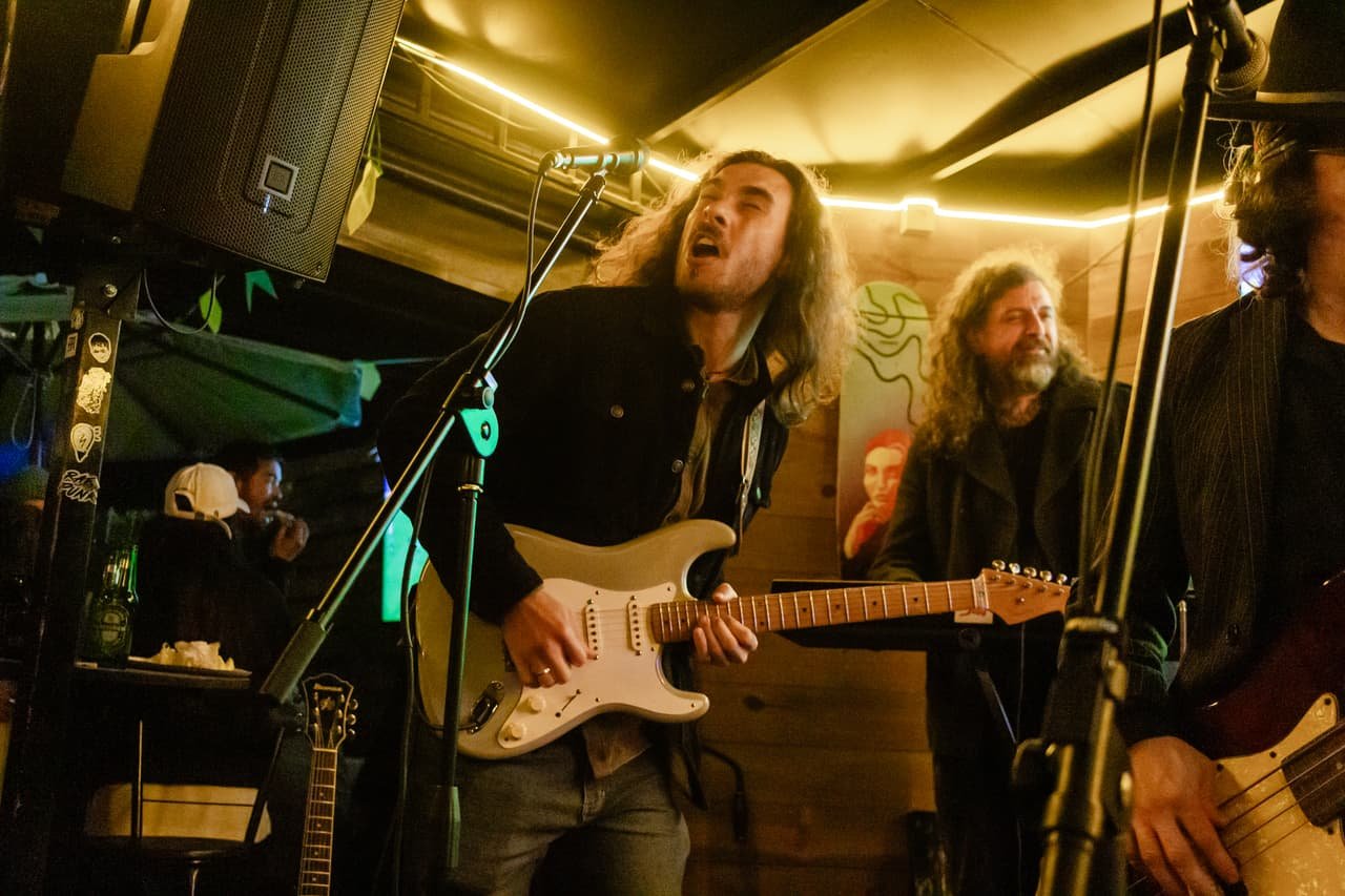 Guitarrista de cabelo comprido sola uma guitarra branca no palco cercado de amplificadores e outros músicos