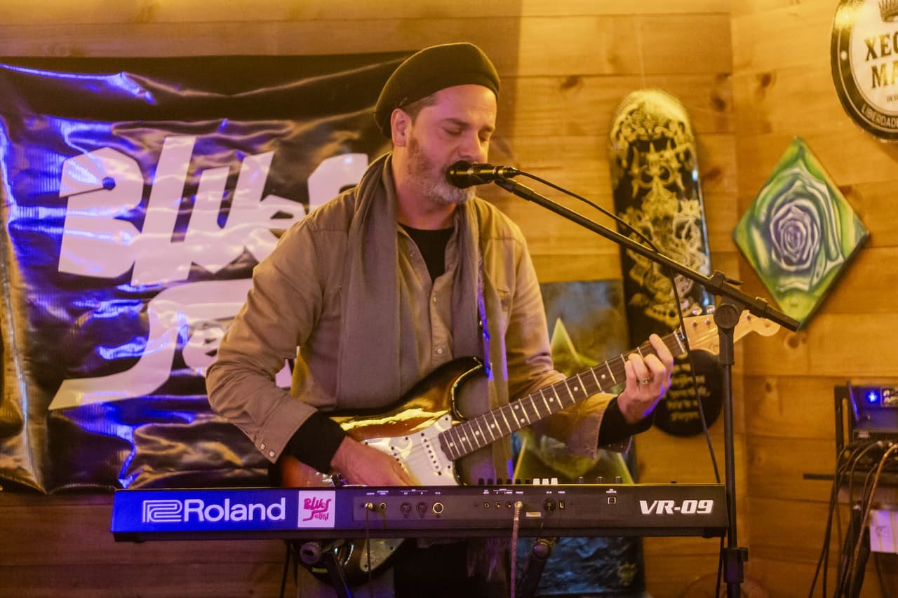 Guitarrista de boina canta e toca guitarra branca em frente ao banner Blues Session e a um teclado Roland