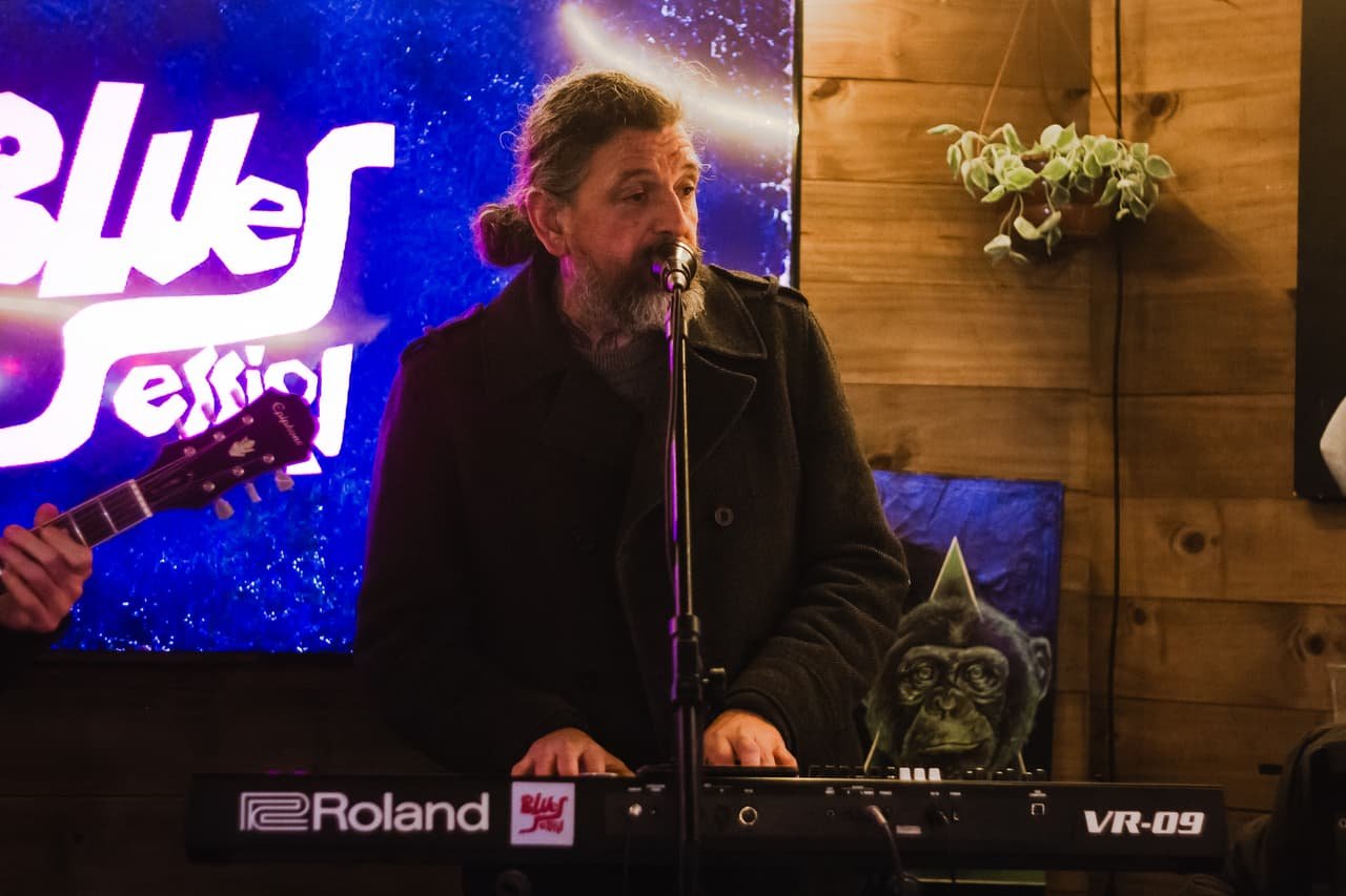 Tecladista canta ao lado do letreiro Blues Session iluminado em azul, sobre parede de madeira