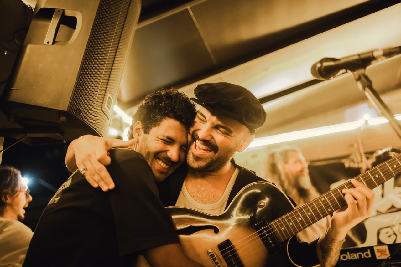 Dois homens se abraçam sorrindo no palco enquanto um deles segura a guitarra, em clima de celebração