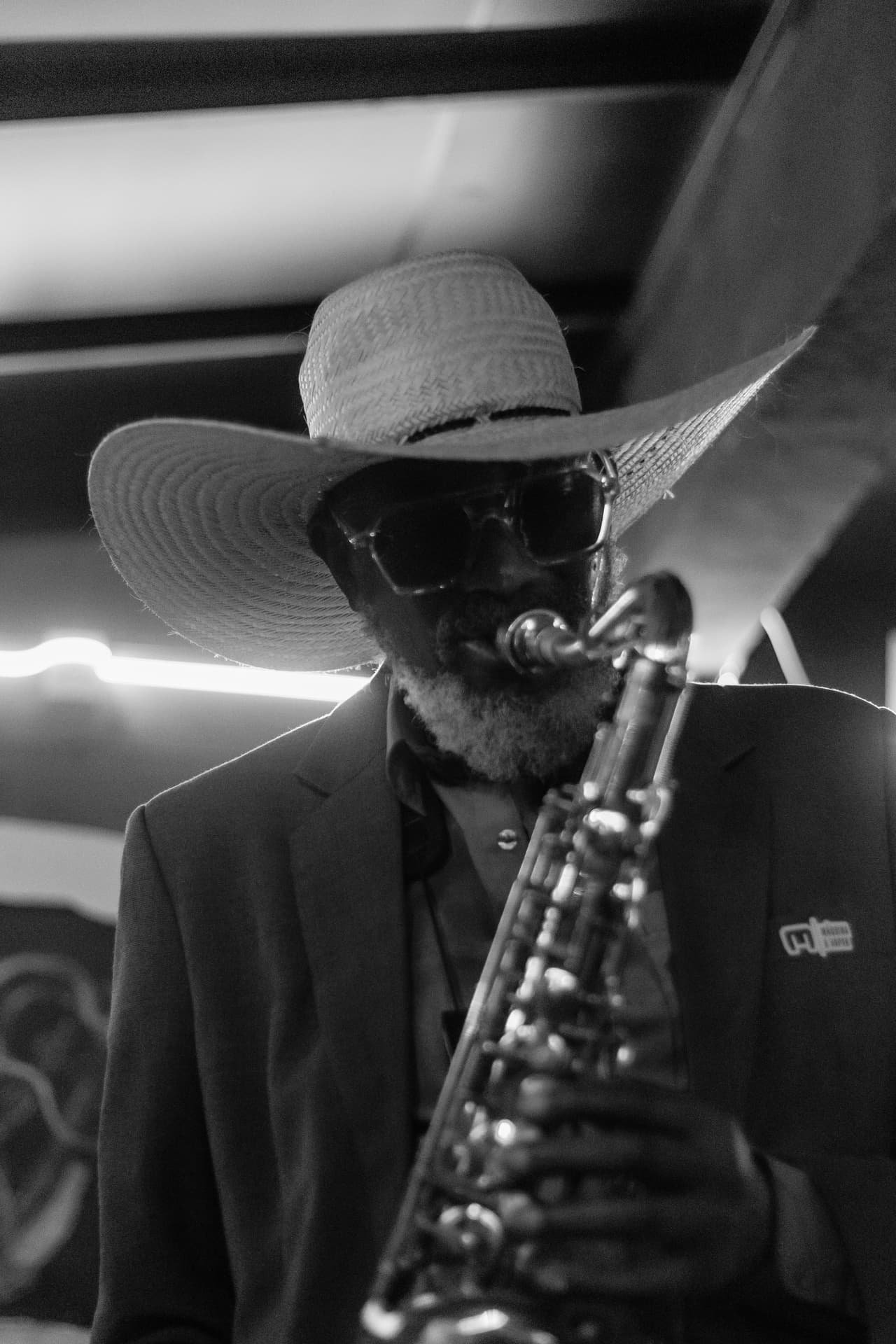 Em preto e branco, saxofonista de chapéu de palha e óculos escuros toca sax de cabeça baixa