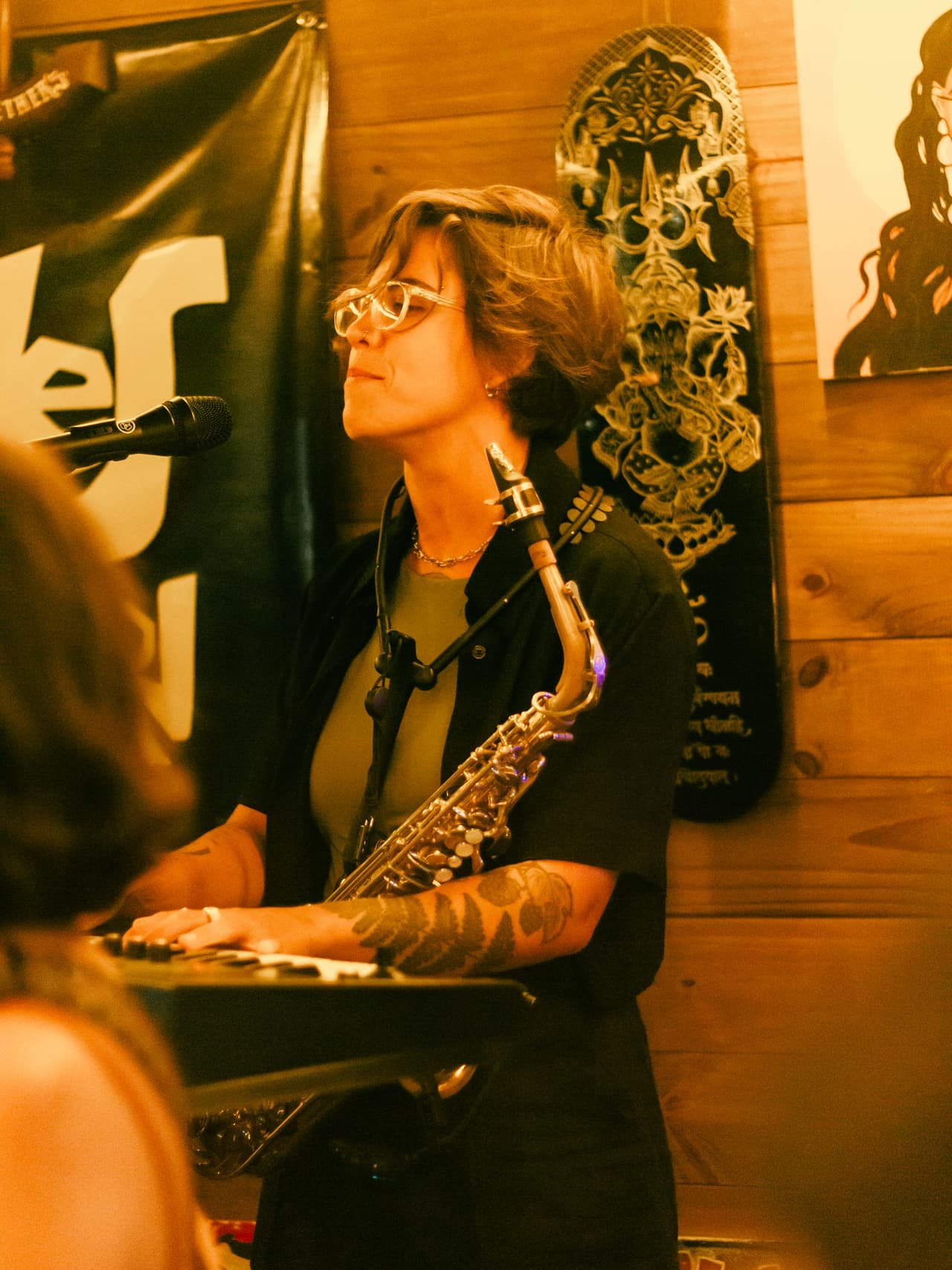 Saxofonista de óculos toca sax alto de olhos fechados diante de um banner na parede de madeira